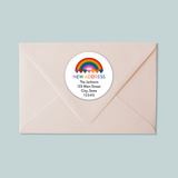 A light pink envelope with a white circular address label featuring a rainbow graphic and text.