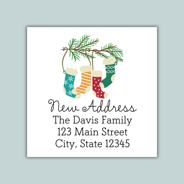 The image is a white envelope with a festive design featuring three colorful Christmas stockings hanging from a branch with pine needles. The text on the envelope reads "New Address The Davis Family 123 Main Street City, State 12345".