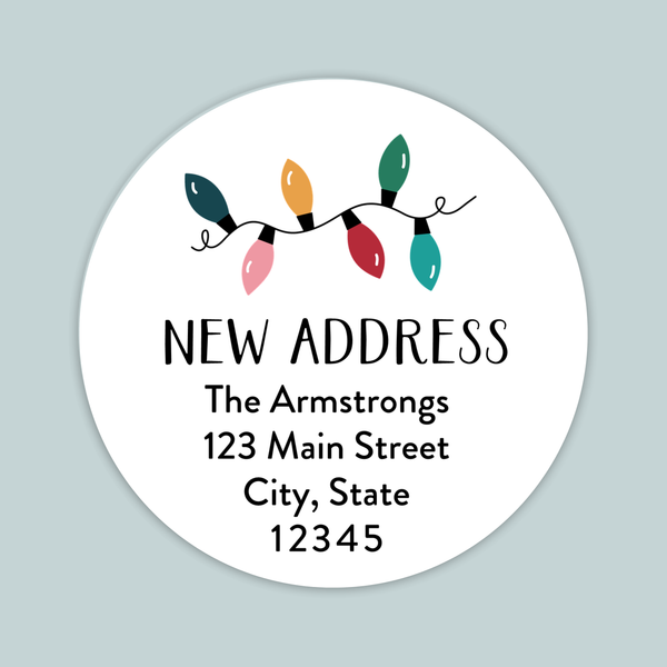 a white circular label with a festive design featuring colorful Christmas lights. The label has text that reads "NEW ADDRESS The Armstrongs 123 Main Street City, State 12345".