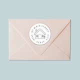 A light pink envelope with a white circular stamp featuring a house illustration and text.